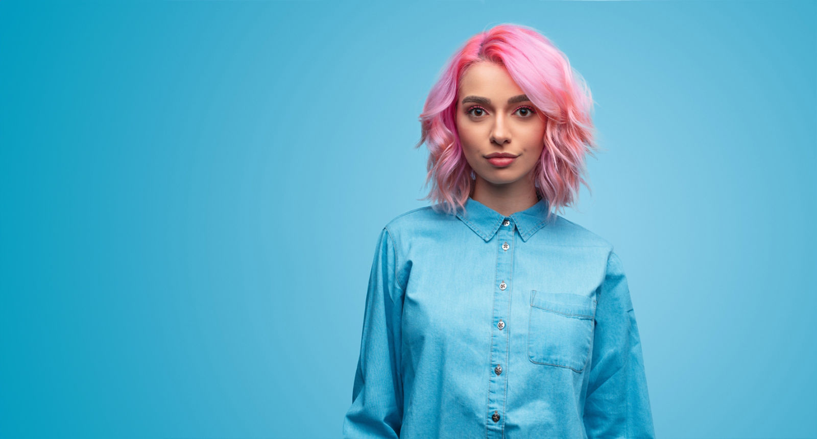 Beautiful young woman with trendy wavy pink hairstyle wearing blue shirt and looking at camera on blue background